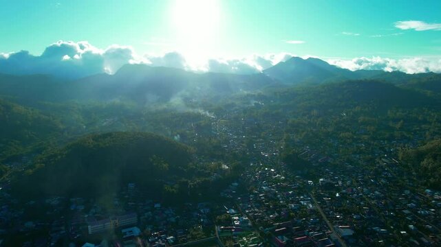 Drone footage showing Bajawa town surrounded by lush green hills and a prominent volcano in the distance under a clear blue sky. Captured during early morning with soft natural light.