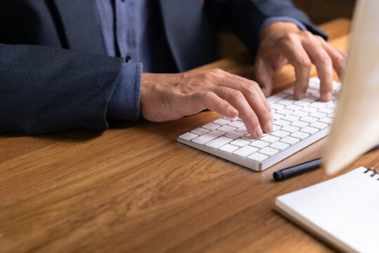 Close-up of hands of a businessman typing on a white wireless keyboard on wooden desk, representing office work, technology, productivity, and modern business communication