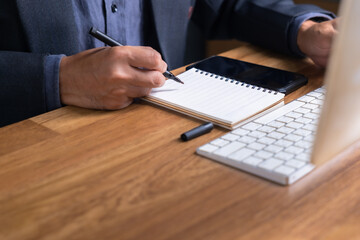 Close-up of businessman’s hands writing in a spiral notebook with a pen beside smartphone and...