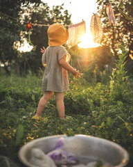 A little girl  in a yellow hat standing in a lush garden during sunset, gazing at hanging clothes...
