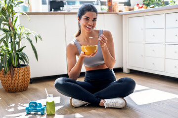 Athletic woman eating a healthy bowl of muesli with fruit sitting on floor in the kitchen at home