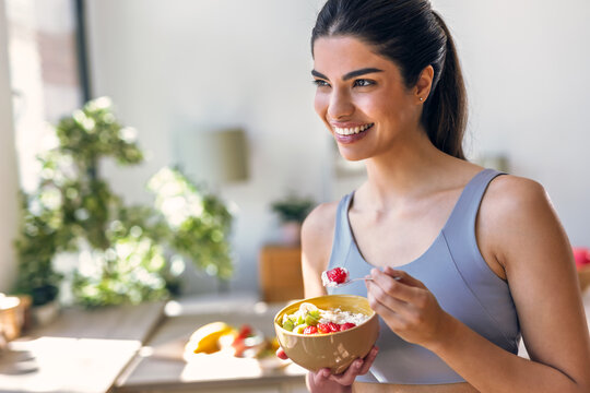 Smiling sporty woman eating a healthy fruit bowl while sitting in the kitchen at home - Powered by Adobe
