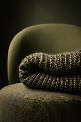 A cozy, rolled knitted blanket in dill green color rests on a soft, rounded chair. The scene conveys warmth and comfort in a minimalist setting.