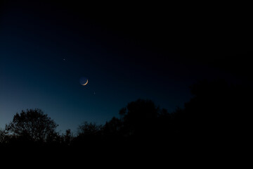Crescent young Moon with stars, planets and countryside silhouettes.