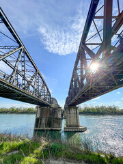 A low-angle view of two robust metal truss bridges spanning the Po River near Cremona, with bright sunlight glinting off the water and a clear blue sky.