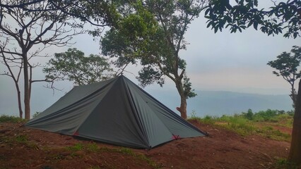 A dark-colored flysheet tent stands on a misty mountain peak, surrounded by tall trees and foggy skies, creating a serene and secluded camping atmosphere.