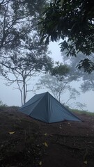 A dark-colored flysheet tent stands on a misty mountain peak, surrounded by tall trees and foggy skies, creating a serene and secluded camping atmosphere.