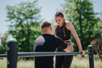 A fitness coach helps a woman perform strength exercises on an outdoor pull-up bar in a sunny park...