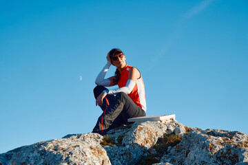 A female hiker enjoys the serene beauty of the mountain landscape, sitting on a rock at the summit,...
