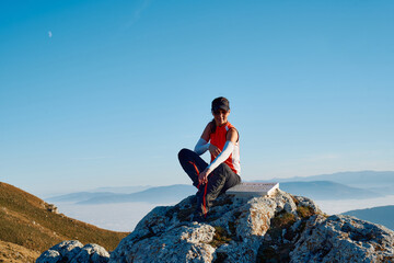 A female hiker enjoys the serene beauty of the mountain landscape, sitting on a rock at the summit,...