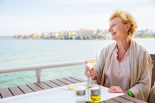 Mature attractive woman traveler sitting alone on the terrace of coffee shop and drinking rose wine. Active life of the elderly in retirement, active seniors - Powered by Adobe