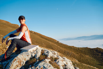 A female hiker enjoys the serene beauty of the mountain landscape, sitting on a rock at the summit,...