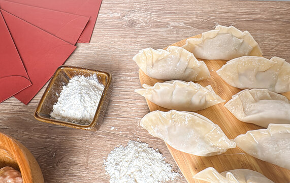 Top-down view of uncooked Chinese dumplings arranged on a bamboo board with flour, dough, and red envelopes. Symbolic of festive food prep for Chinese New Year celebration.
- Powered by Adobe