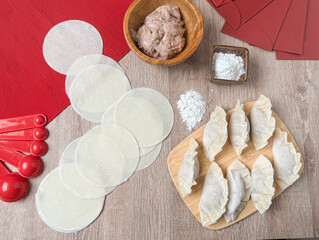 Top-down view of Chinese dumpling preparation with wrappers, pork filling, flour, bamboo board, and red envelopes. Includes raw jiaozi and festive elements for Chinese New Year cooking.