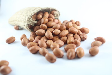 A rustic burlap sack overflows with peanuts onto a clean white surface, highlighting natural,...