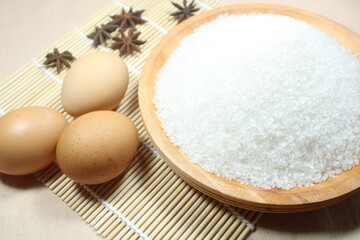 A rustic kitchen scene featuring brown eggs, a wooden bowl of granulated sugar, and star anise pods, all arranged on a natural bamboo mat
