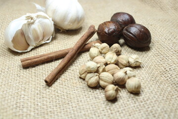 A rustic assortment of whole garlic, nutmegs, cinnamon sticks, and cardamom pods arranged on a...