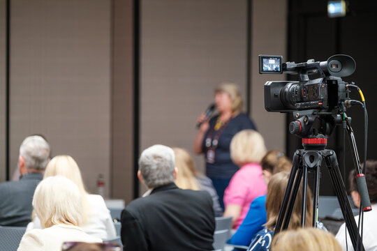 Video camera capturing live speaker at business conference with audience in attendance.