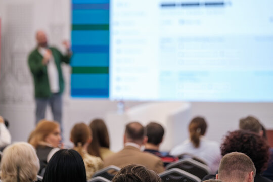 Speaker presenting information to audience at conference with blurry projection screen visible.