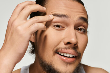 Handsome young bearded man smiling while touching his eyebrow in bright studio setting