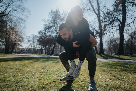 A playful and joyful moment as a young man carries a woman on his back in a sunny park, showcasing energy and happiness.