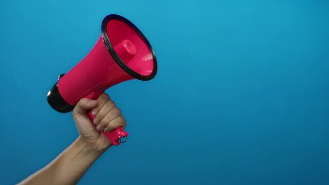 Hand holding a pink megaphone against a solid blue wall background, emphasizing communication and announcement themes.