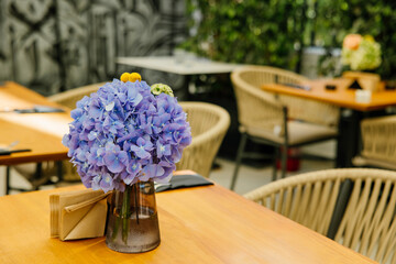 Vase with purple hydrangeas and yellow billy balls placed on a wooden table in a stylish outdoor café setting.

