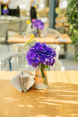 Bright cafe scene with a glass vase of purple hydrangeas and yellow billy balls placed on a wooden table in sunlight.
