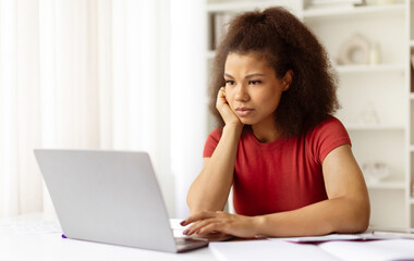 Frustrated young black woman staring at laptop with hand on face during remote work or study....