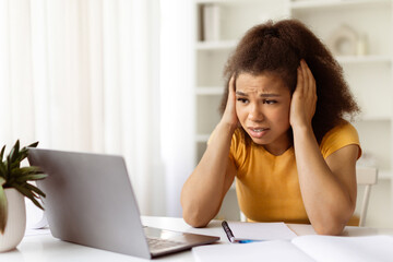 Stressed young black woman sitting at home desk with hands on head, worried while looking at...