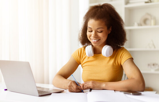 Young black woman with headphones around neck smiling while writing in notebook during remote study. Peaceful home workspace, focus and productivity in warm lighting, copy space