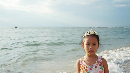Young girl in a floral dress and tiara stands on a beach shore with gentle waves and cloudy sky - Powered by Adobe