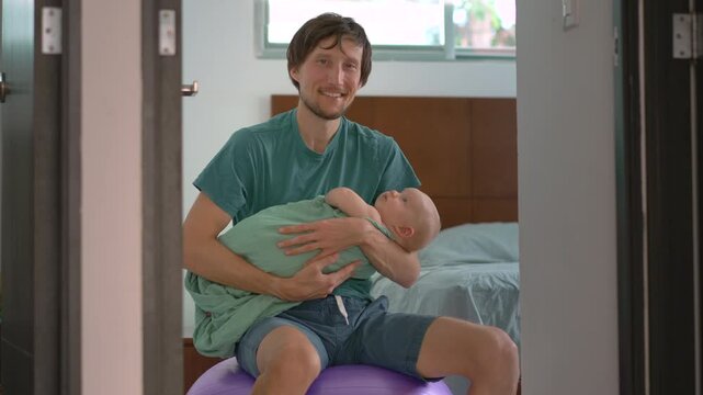 Loving father rocks his swaddled newborn baby to sleep while gently bouncing on a fitball, showing tender bonding, parental care, and soothing bedtime routine at home. Slowmotion video.