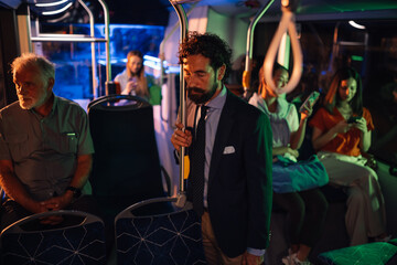 Businessman standing on bus at night holding handrail looking tired after work
