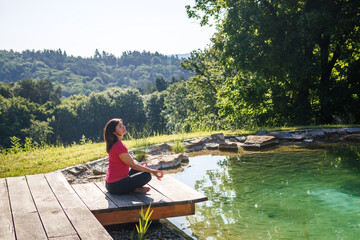 Mature woman meditating in yoga lotus position by natural swimming pond in backyard garden. Healthy...