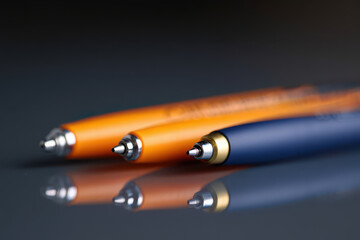 Close up shot of two orange pens and one blue pen on a reflective surface with a dark background