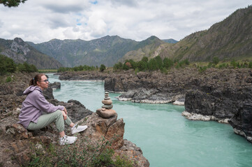 A woman in casual outdoor clothing sits on a hillside, admiring a breathtaking view of mountains, a winding Katun river. Trip on Altai Mountains in Altai Republic