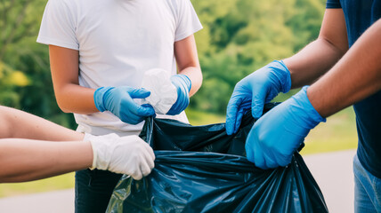 A close-up photograph of a community cleanup activity. Two people wearing blue latex gloves are sorting waste into a large black garbage bag.