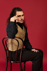 Captivating young man in stylish vest sitting thoughtfully on a chair against a vibrant red backdrop