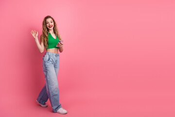 Cheerful young woman in stylish outfit waving hello against pink background while holding a phone...