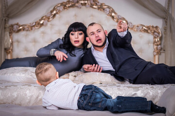 A two year old son sits on the edge of the bed. Dad and pregnant mom are watching him from behind....