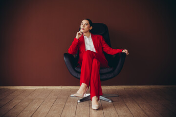 Attractive Confident Businesswoman in Red Suit Sitting in Stylish Chair in Brown Office Setting