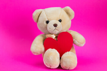 White toy cartoon teddy bear sits on a pink background with red paper heart
