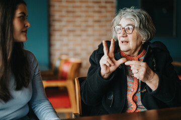 An older woman in glasses converses expressively with a younger seated woman in a cozy indoor environment.