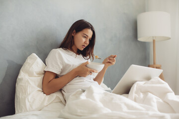 Young Woman Eating Breakfast and Working on Laptop in Bed with Cozy Light