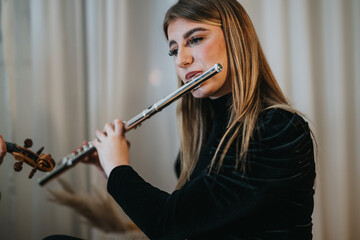 A young woman is seen playing a shiny flute in a softly lit interior setting. Her focused...