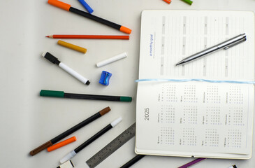 A flat lay of a 2025 calendar planner with a monthly goal page, surrounded by pens, markers, ruler, sharpener, and colored pencils on a white desk