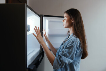 Young woman looking inside an empty refrigerator, expressing disappointment and concern about food...