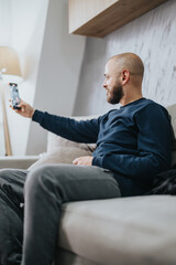 A man with a beard sitting on a comfortable sofa indoors, capturing a selfie with his smart phone,...