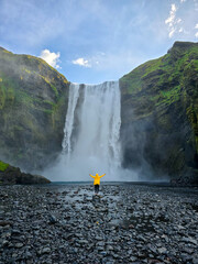 A vibrant traveler wearing a yellow jacket stands in awe before a stunning waterfall in Iceland,...